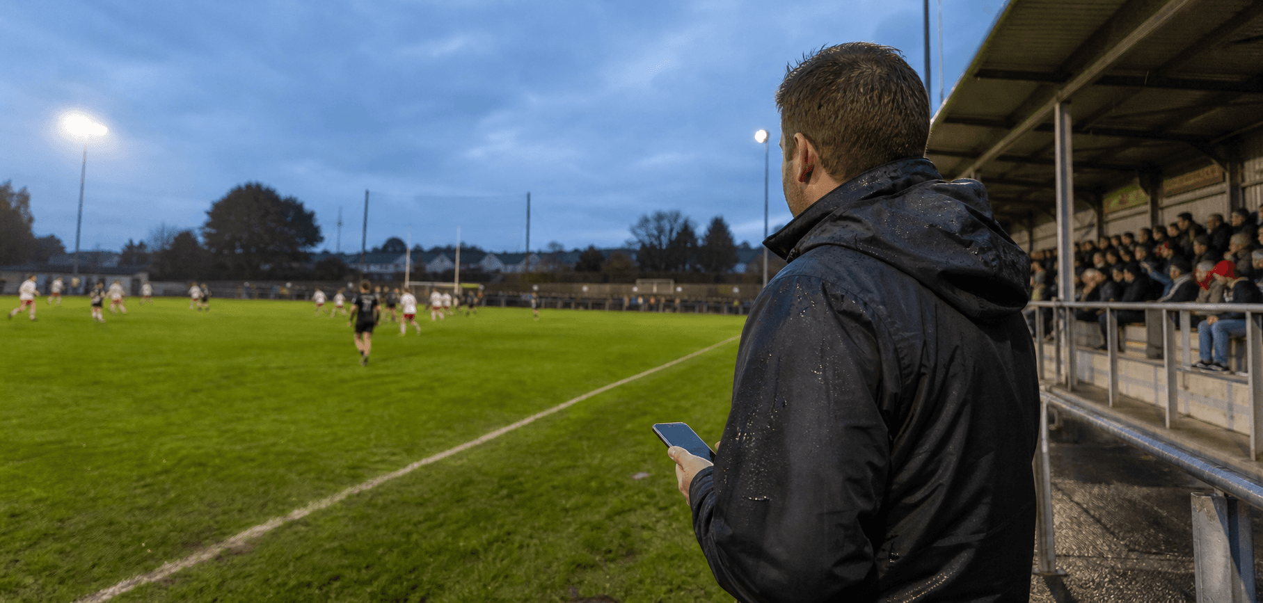 A sideline reporter publishing from a phone during an evening Gaelic football match at a local Irish ground
